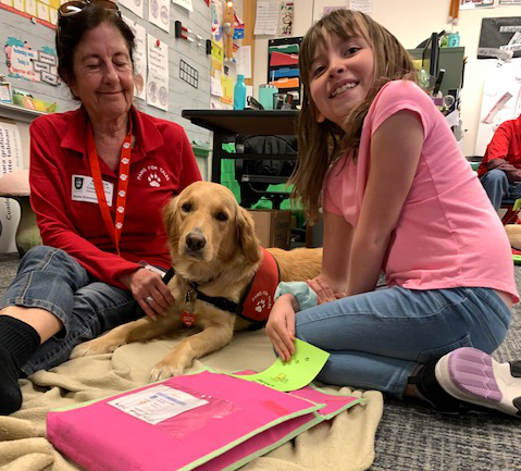 A smiling student with a Paws for Tales Volunteer and her therapy dog.